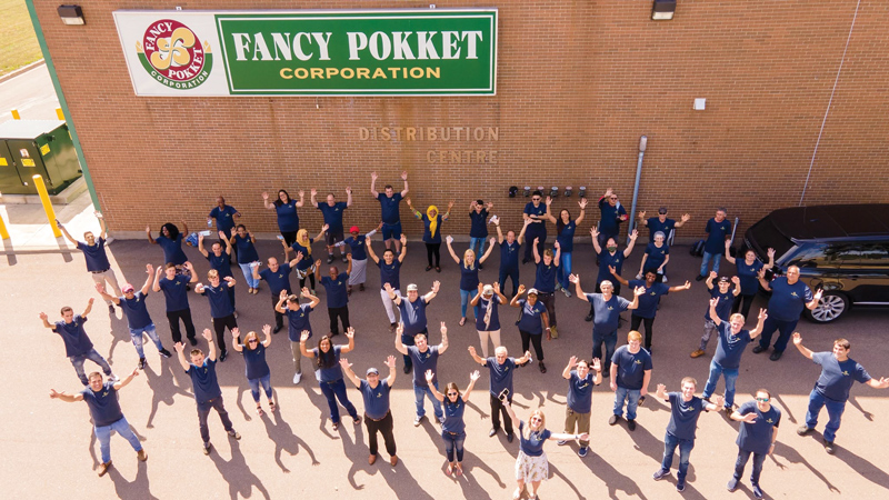 a large group of people dressed in dark coloured t shirts are standing, spaced apart, and with their arms in the air. They are outside a large building, and it is a sunny day.