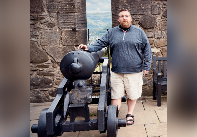 a man wearing khaki shorts and a long sleeve sweatshirt is standing with his left hand in his pocket and his right hand leaning on a canon (or replica of a canon) in a space with stone 'tile' floor and rough large stone wall. it may be a museum of some type.