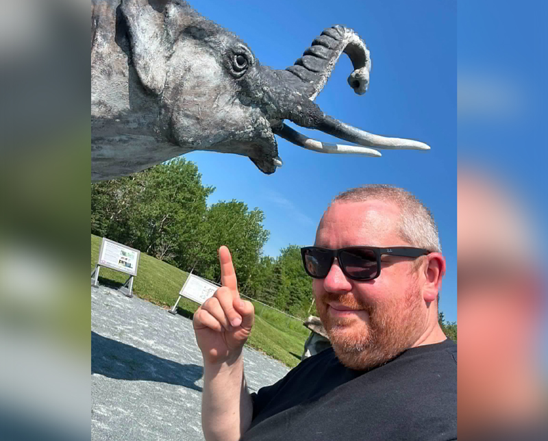 a man, lower right, is shown from the shoulders up, and pointing upwards towards an elephant statue in the upper right. The background is a park parking lot it seems, and the sky is blue.