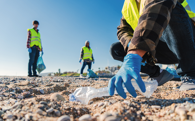 3 workers wearing yellow safety vests and latex gloves are on a beach gathering trash . It is a view from the ground, and the worker at the right is very close, and you can see the hand picking up a disgarded plastic bottle.