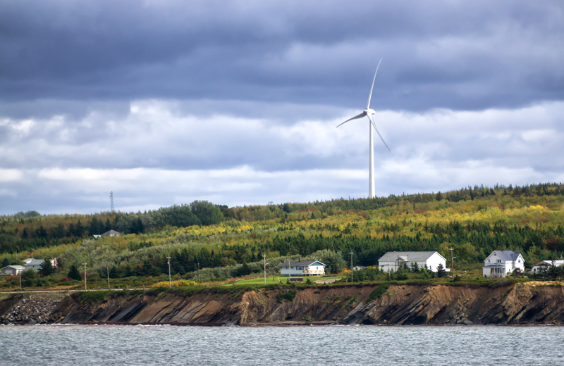 a Wind turbine is visible over a low hill behind a small community on the sea coast. The hill is wooded, and the sky is cloudy and a little dark.